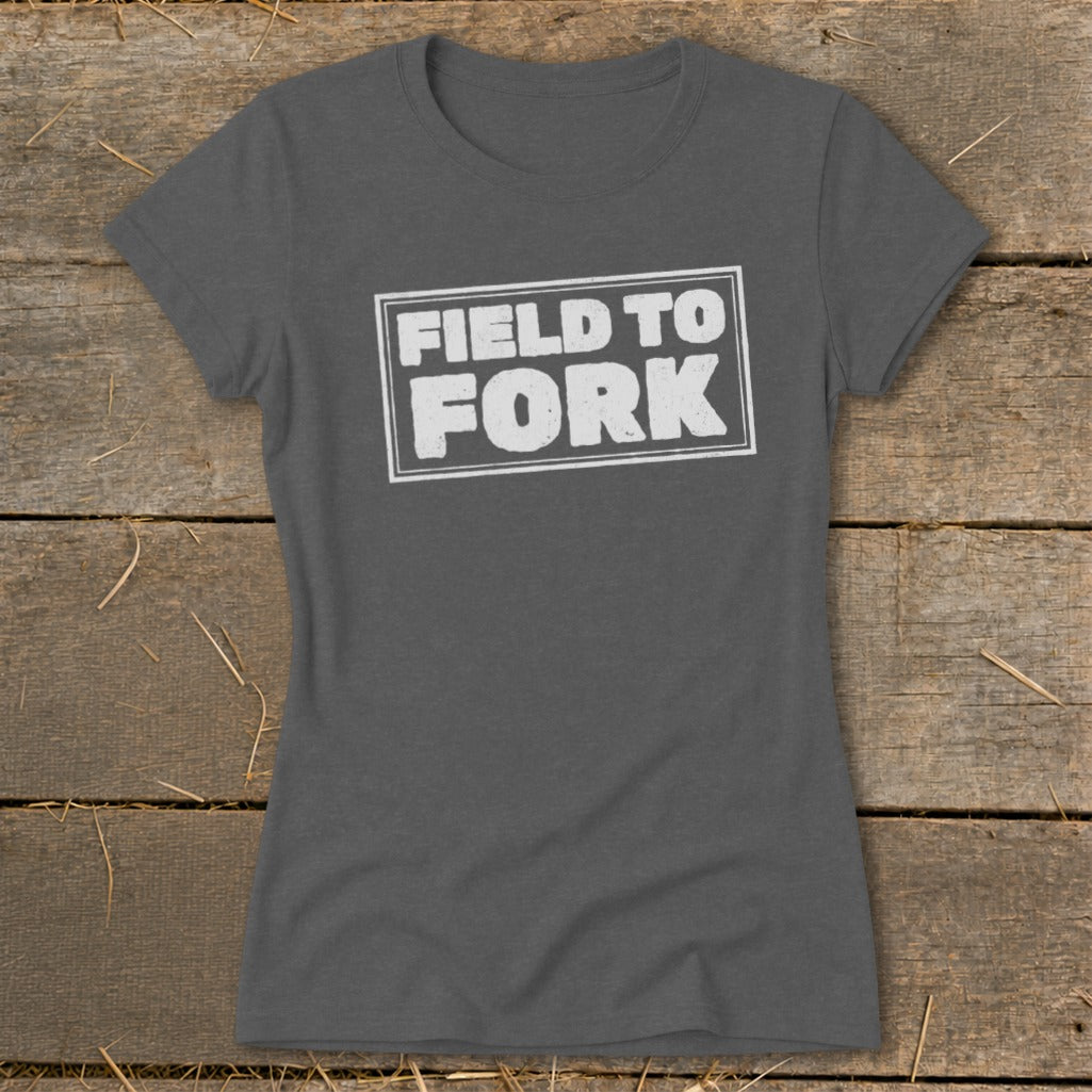 Dark grey women's t-shirt featuring the white text Field to Fork inside a bordered rectangle, laid on a rustic wooden background.