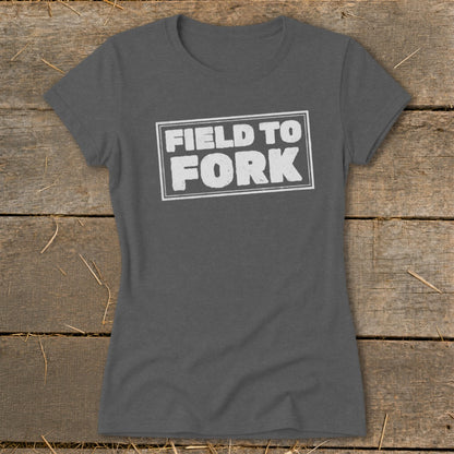 Dark grey women's t-shirt featuring the white text Field to Fork inside a bordered rectangle, laid on a rustic wooden background.