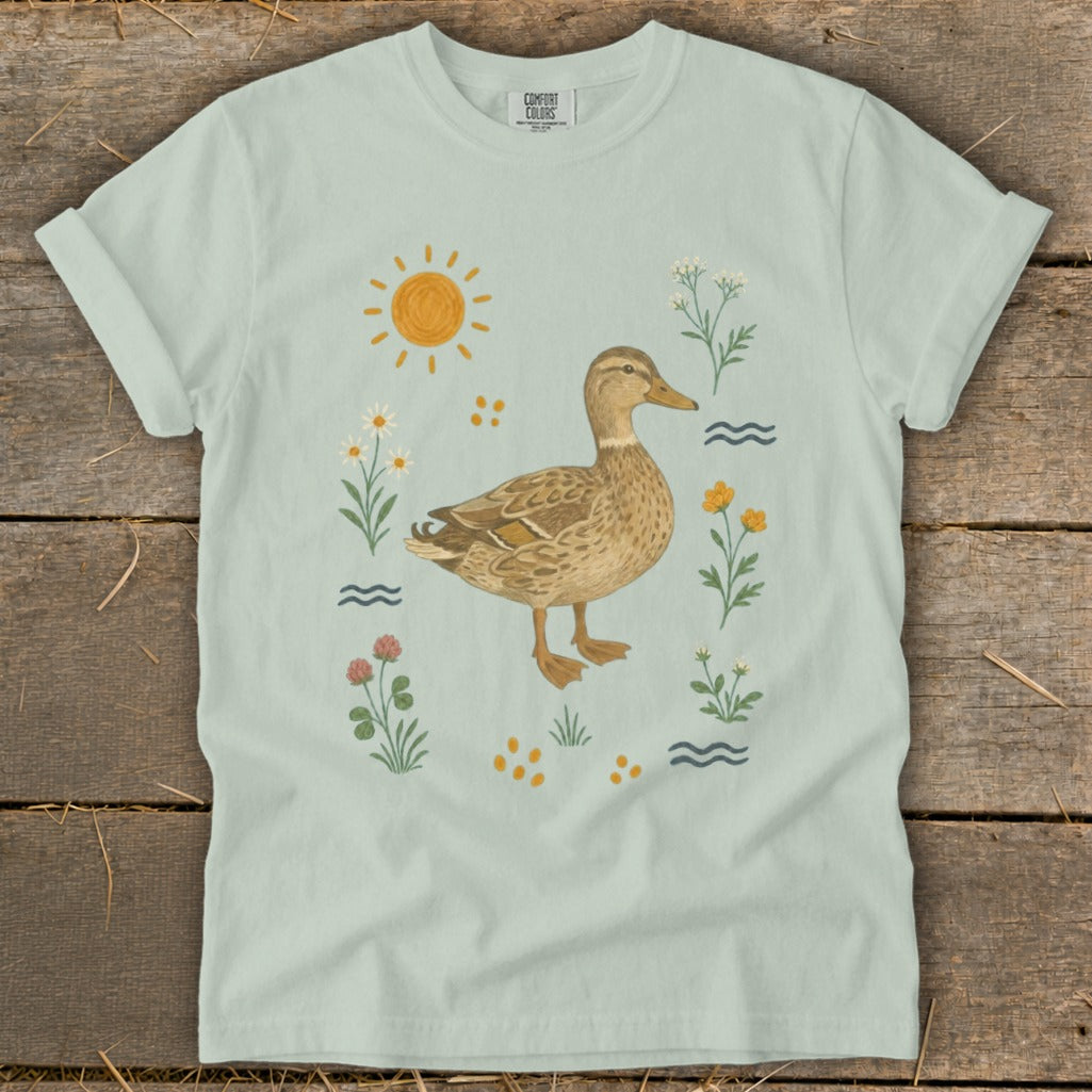 Light blue t-shirt with a duck illustration surrounded by flowers, sun, and water waves.