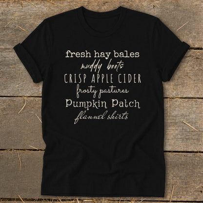 Black t-shirt with white text listing fall themes: fresh hay bales, muddy boots, crisp apple cider.