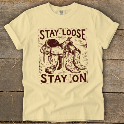 Yellow t-shirt with maroon graphic of a saddle and cowboy boots, saying "STAY LOOSE STAY ON".