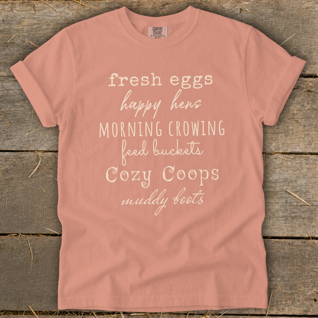 Peach t-shirt with text: fresh eggs, happy hens, morning crowing, feed buckets, cozy coops, muddy boots.