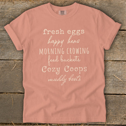 Peach t-shirt with text: fresh eggs, happy hens, morning crowing, feed buckets, cozy coops, muddy boots.