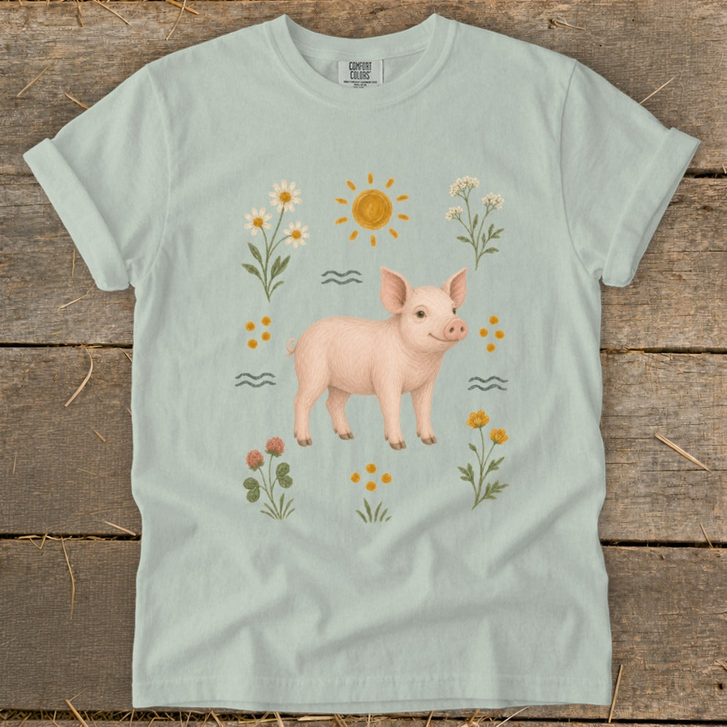 Light blue t-shirt with a pink pig illustration surrounded by flowers and a sun.