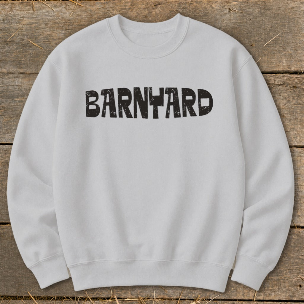 A light gray crewneck sweatshirt laid flat on a wooden background, featuring the distressed black text "BARNYARD" centered on the chest.