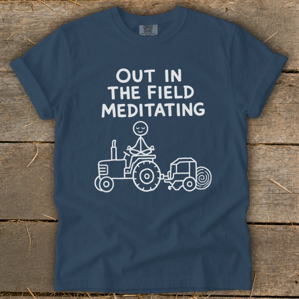 Navy t-shirt with white text "OUT IN THE FIELD MEDITATING" above a stick figure meditating on a tractor.
