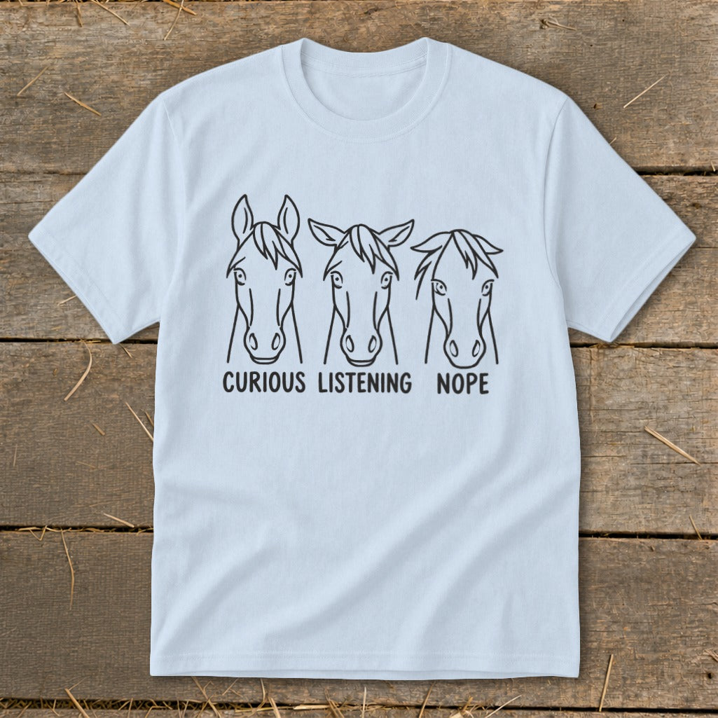 Light blue t-shirt featuring three horse face illustrations labeled Curious, Listening, and Nope, laid flat on wooden planks.