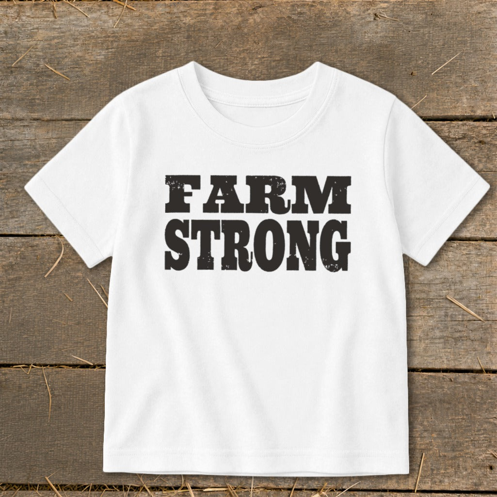 A white children's t-shirt printed with the text "FARM STRONG" in distressed black letters lies flat on a wooden background.