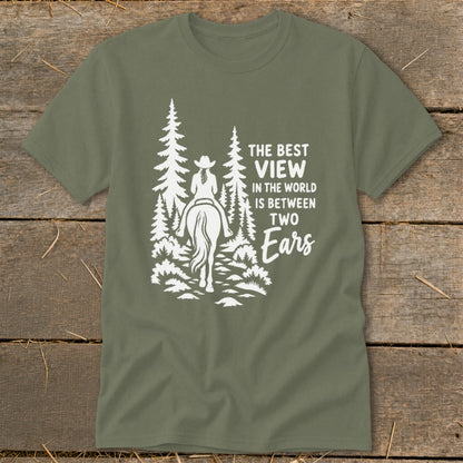 An olive green t-shirt with white graphic text reading The Best View in the World is Between Two Ears alongside a horse rider in a forest.