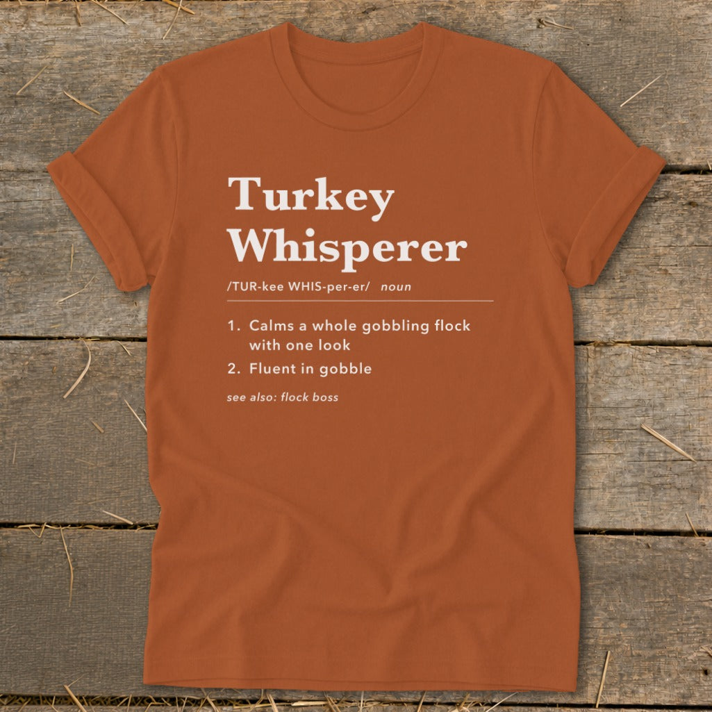 Orange t-shirt with "Turkey Whisperer" definition: "Calms a whole gobbling flock," "Fluent in gobble."