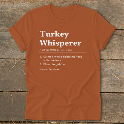 Orange t-shirt with "Turkey Whisperer" definition: "Calms a whole gobbling flock," "Fluent in gobble."