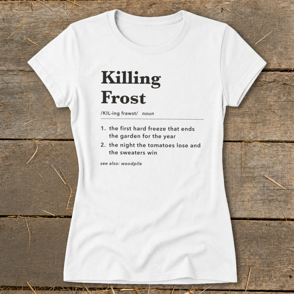A white short-sleeved t-shirt lies flat on a wooden surface, featuring black text defining "Killing Frost."