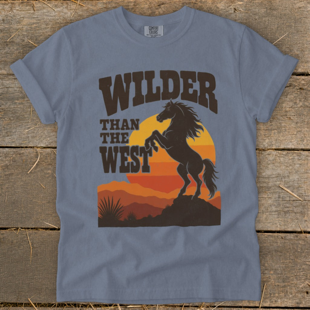T-shirt design with dark brown text "WILDER THAN THE WEST" above a rearing horse silhouette at sunset.