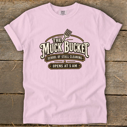 Pink t-shirt with "The Muck Bucket" logo, "School of Stall Cleaning," and "Opens at 5 AM."