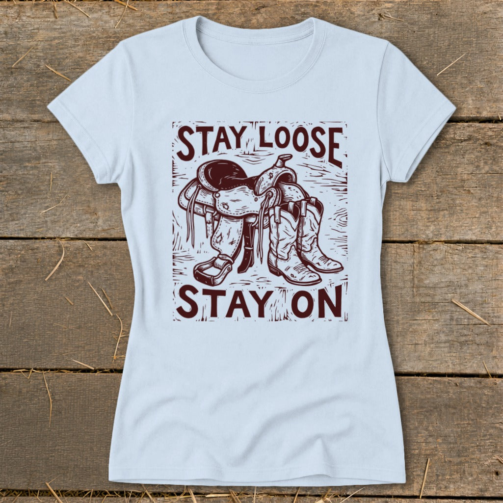 Light blue t-shirt with a graphic of a saddle and cowboy boots and text "STAY LOOSE STAY ON".