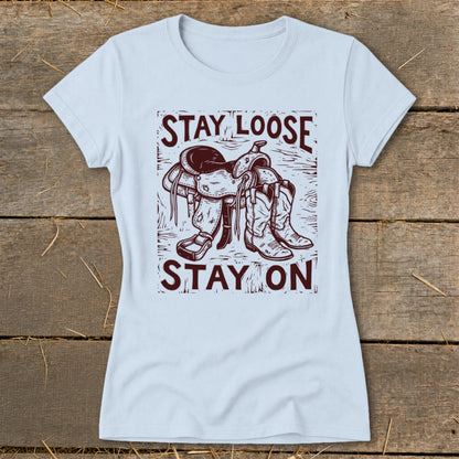 Light blue t-shirt with a graphic of a saddle and cowboy boots and text "STAY LOOSE STAY ON".
