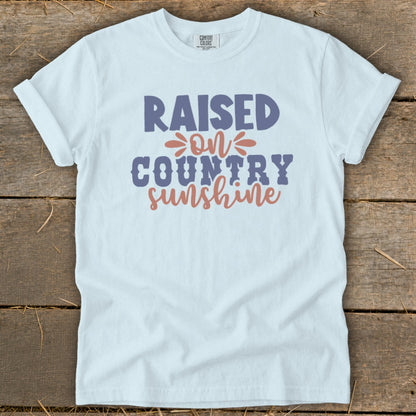 White t-shirt with text "RAISED ON COUNTRY sunshine" in purple and coral.