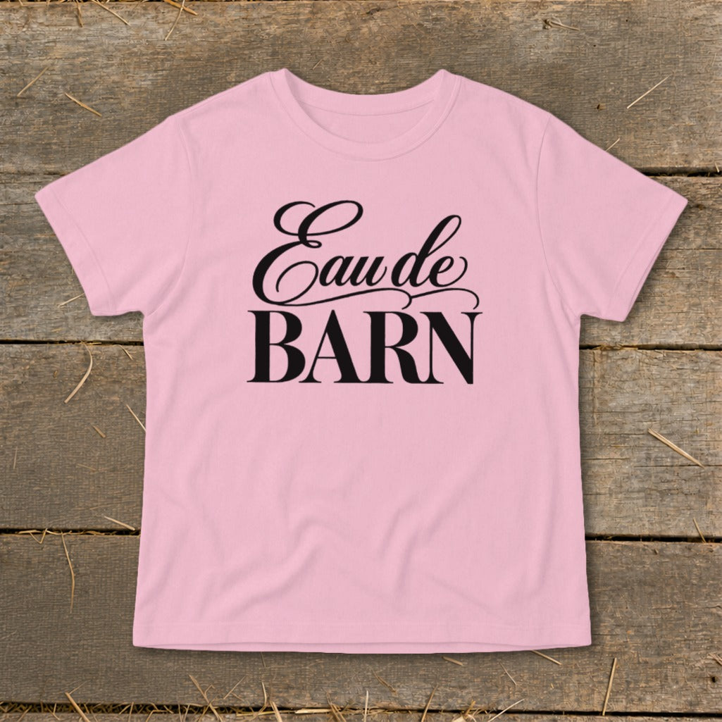 Pink t-shirt with black text "Eau de BARN" on a wooden background.