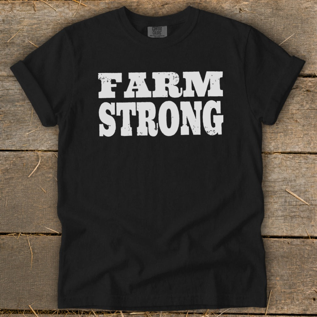 A black t-shirt with rolled-up sleeves lies flat on a rustic wooden surface, displaying white text that reads FARM STRONG.