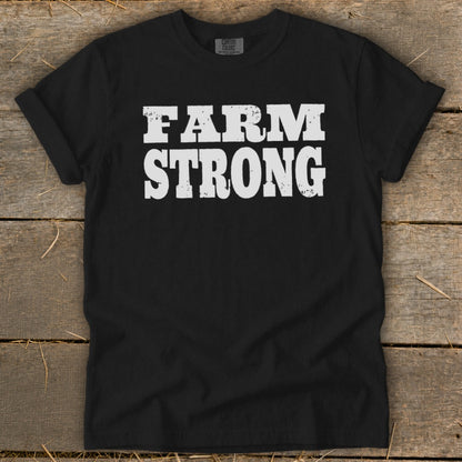 A black t-shirt with rolled-up sleeves lies flat on a rustic wooden surface, displaying white text that reads FARM STRONG.