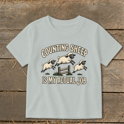 Light blue t-shirt graphic with sheep jumping over a fence and text "COUNTING SHEEP IS MY ACTUAL JOB".