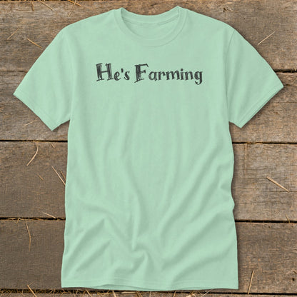 Light green t-shirt with "He's Farming" text in dark grey, on a wooden background.