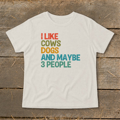 A white t-shirt laid flat on a wooden surface, featuring multicolored distressed text that reads I LIKE COWS DOGS AND MAYBE 3 PEOPLE.