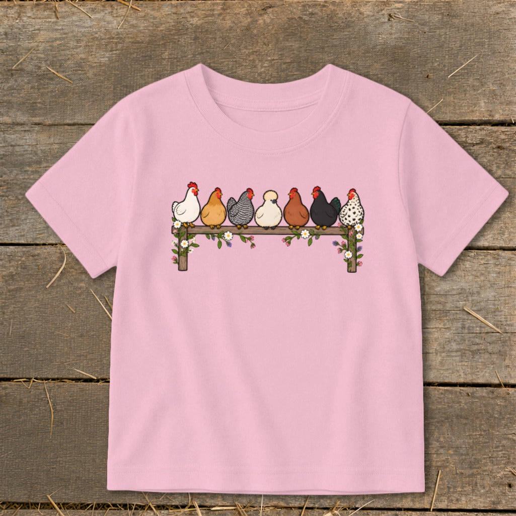 A pink short-sleeved t-shirt features an illustration of seven colorful chickens perched on a wooden fence adorned with flowers.