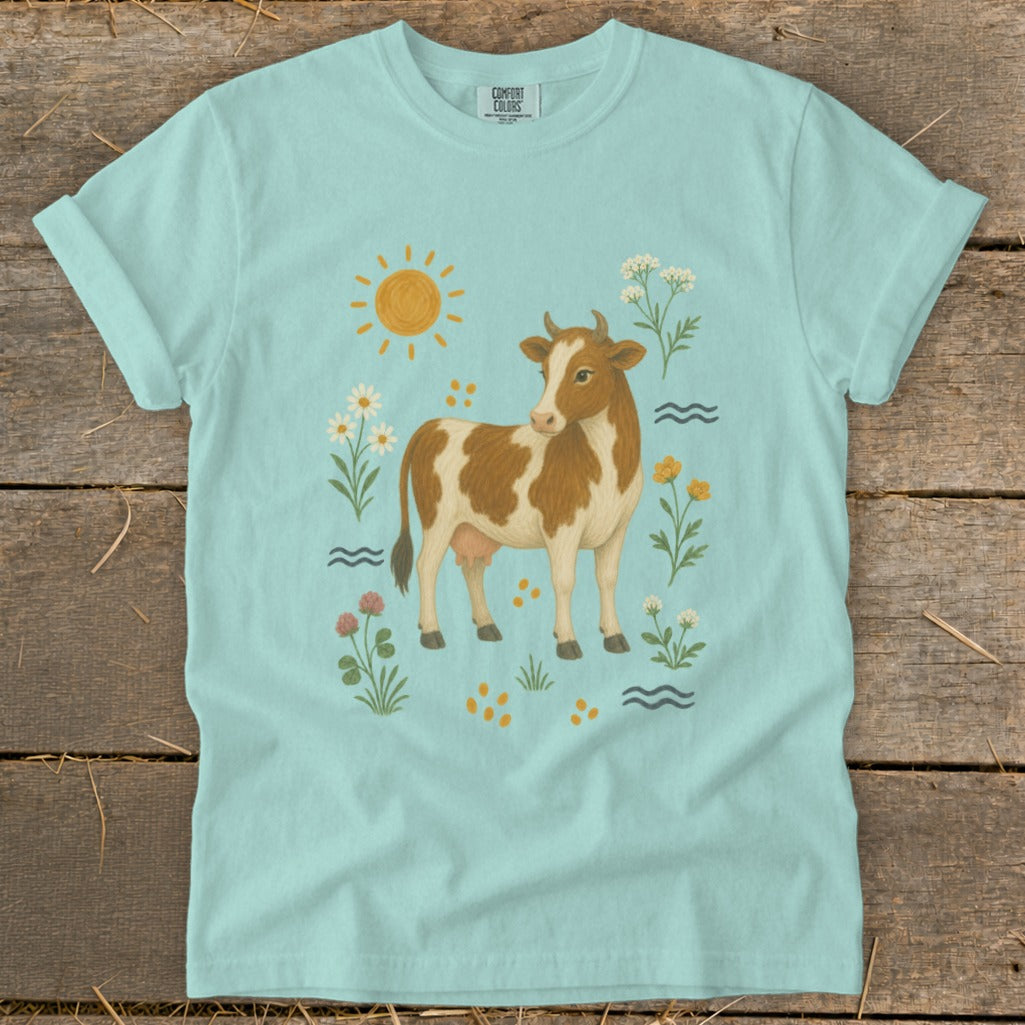 Light blue t-shirt with a brown and white cow surrounded by flowers, sun, and waves.