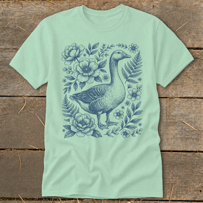 Light green t-shirt with a blue line drawing of a goose surrounded by flowers and ferns.