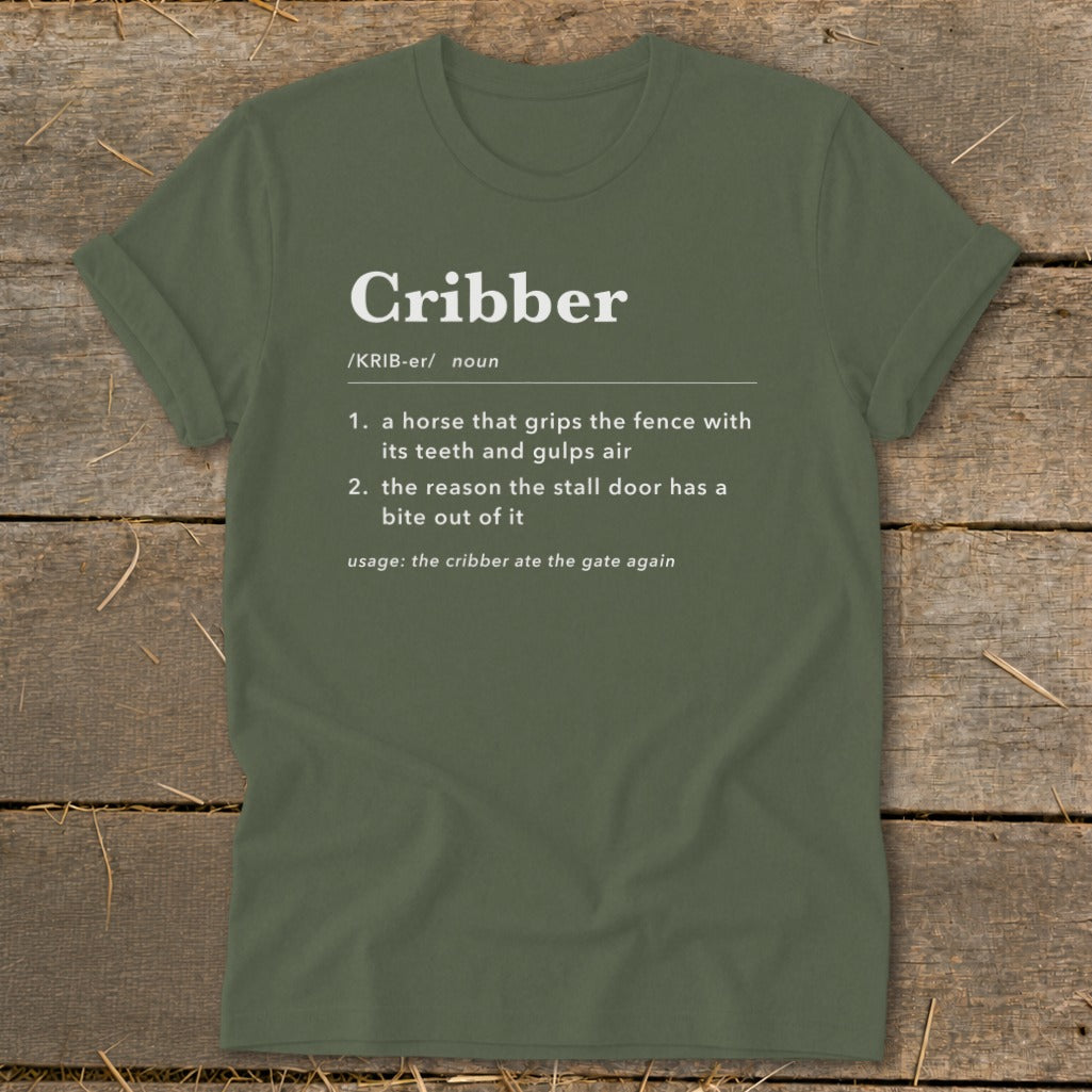 An olive green t-shirt laid flat on a wooden surface, featuring dictionary definitions of the word Cribber in white text.