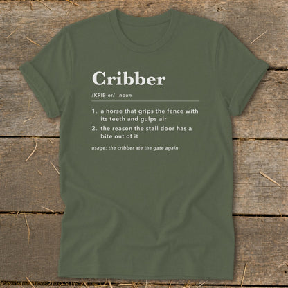 An olive green t-shirt laid flat on a wooden surface, featuring dictionary definitions of the word Cribber in white text.