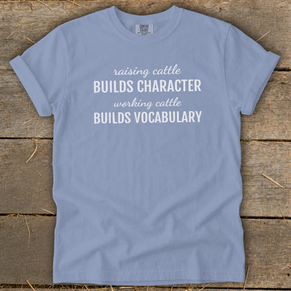 Light blue t-shirt with white text reading "raising cattle BUILDS CHARACTER, working cattle BUILDS VOCABULARY."