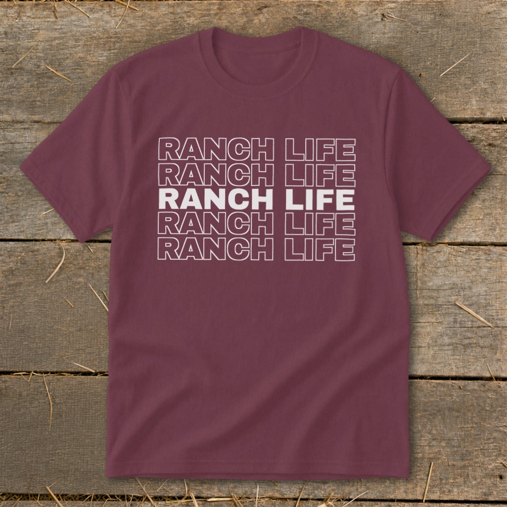 Maroon short-sleeved t-shirt with five rows of stacked white text that reads RANCH LIFE, laid flat on rustic wood.