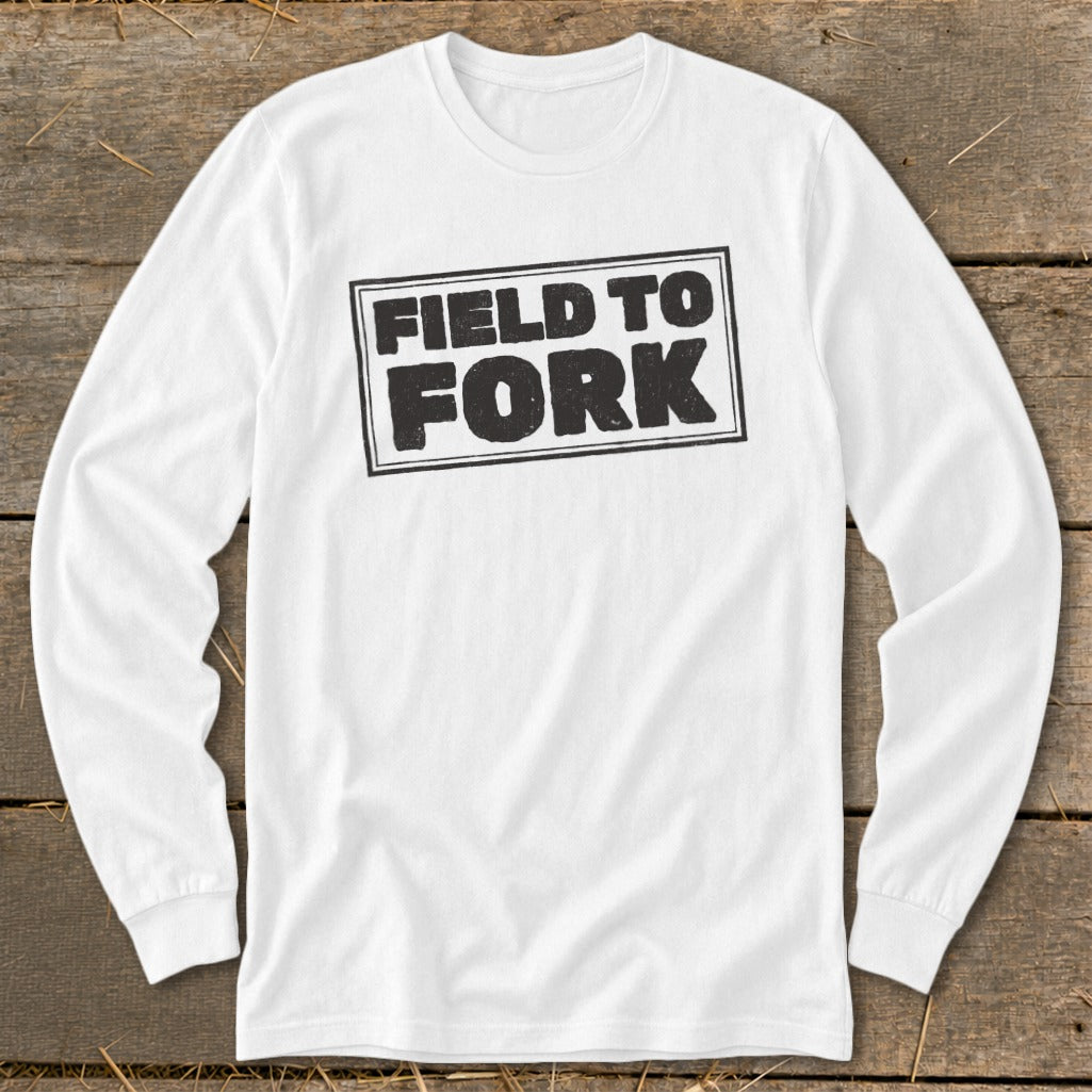 White long-sleeve t-shirt laid flat on wooden background, featuring bold black text reading "FIELD TO FORK" inside a double rectangle.