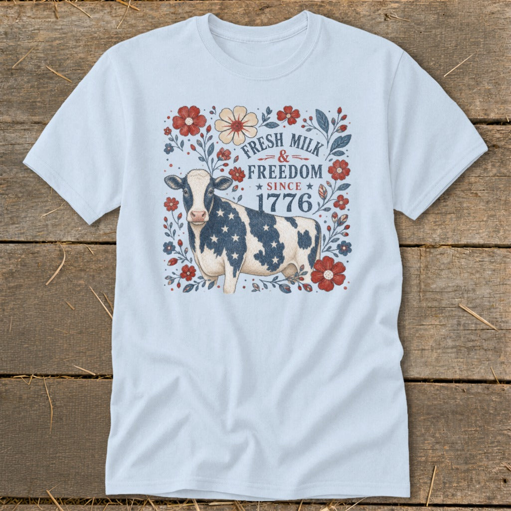 Light blue t-shirt with a cow adorned with stars and flowers, text reads "Fresh Milk & Freedom Since 1776".