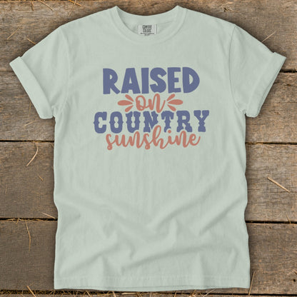 Light green t-shirt with "Raised on Country Sunshine" text in blue and coral.