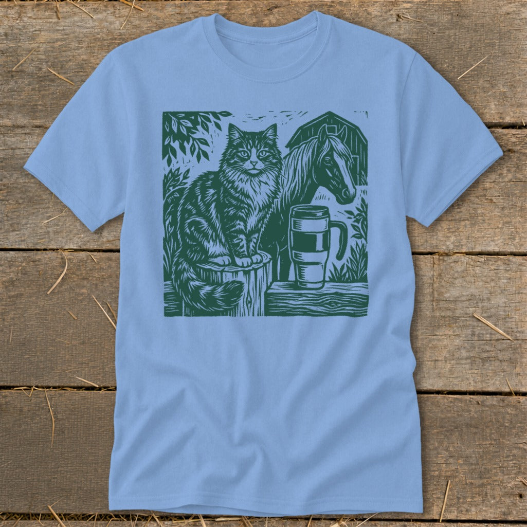 Green linocut design of cat, horse, mug, and barn on light blue t-shirt.