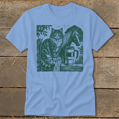 Green linocut design of cat, horse, mug, and barn on light blue t-shirt.