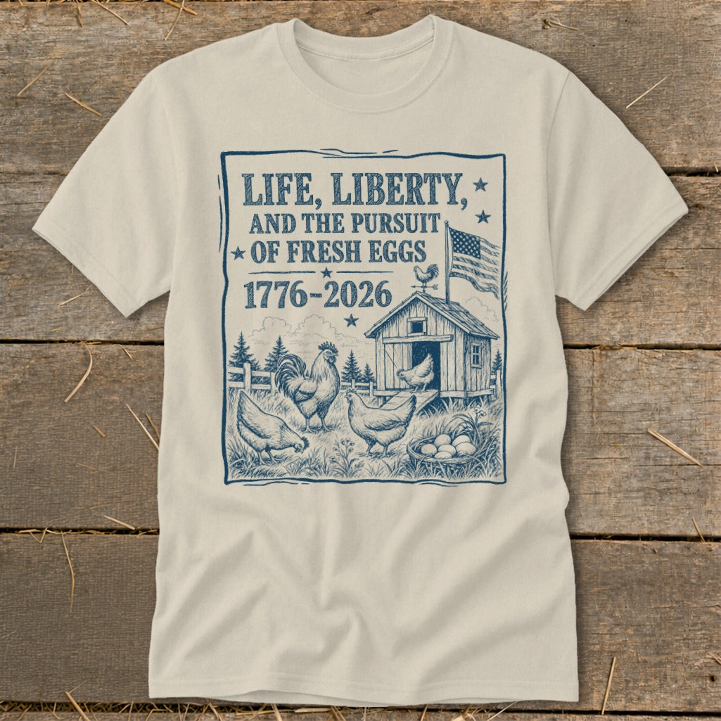Cream t-shirt with a blue graphic of chickens, a coop, and American flag. Text reads "Life, Liberty, and the Pursuit of Fresh Eggs 1776-2026".