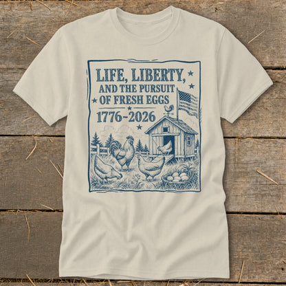 Cream t-shirt with a blue graphic of chickens, a coop, and American flag. Text reads "Life, Liberty, and the Pursuit of Fresh Eggs 1776-2026".