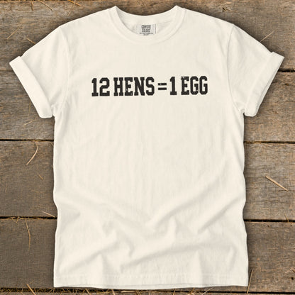 Off-white t-shirt with rolled sleeves displaying black block text reading 12 HENS = 1 EGG on a wooden background.