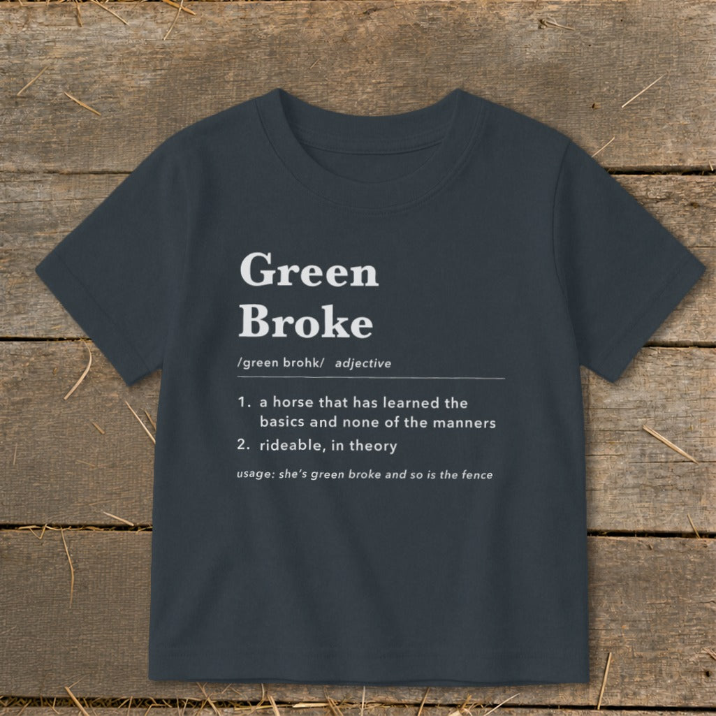 Dark gray short-sleeved children's t-shirt printed with a humorous definition of Green Broke against a rustic wooden background.