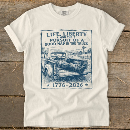 White t-shirt with a blue graphic of a man napping in a truck and text "Life, Liberty and the pursuit of a good nap in the truck 1776-2026".