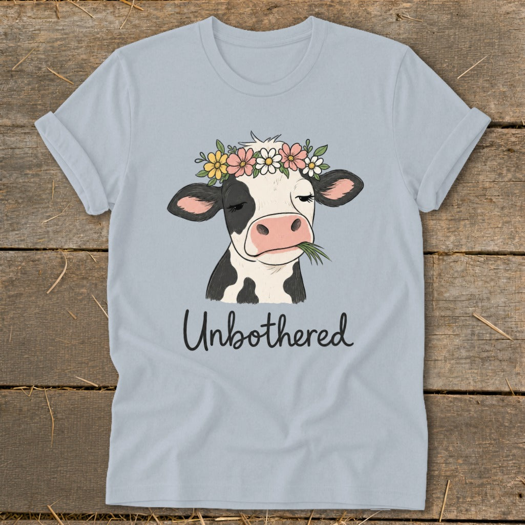 Light blue t-shirt with a cartoon cow wearing a flower crown, chewing grass, and text that reads "Unbothered".