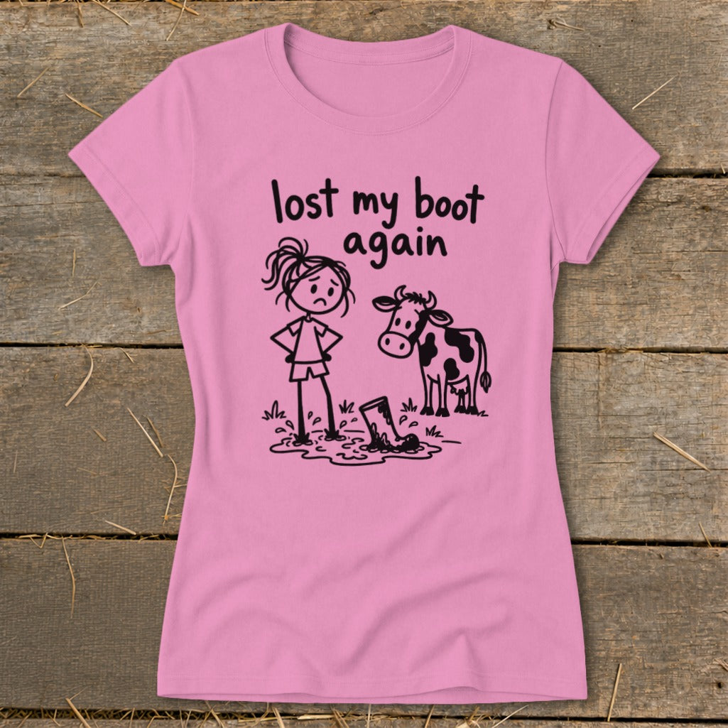 A pink short-sleeved t-shirt features a black cartoon graphic of a sad girl and a cow looking at a muddy boot, with the text "lost my boot again."