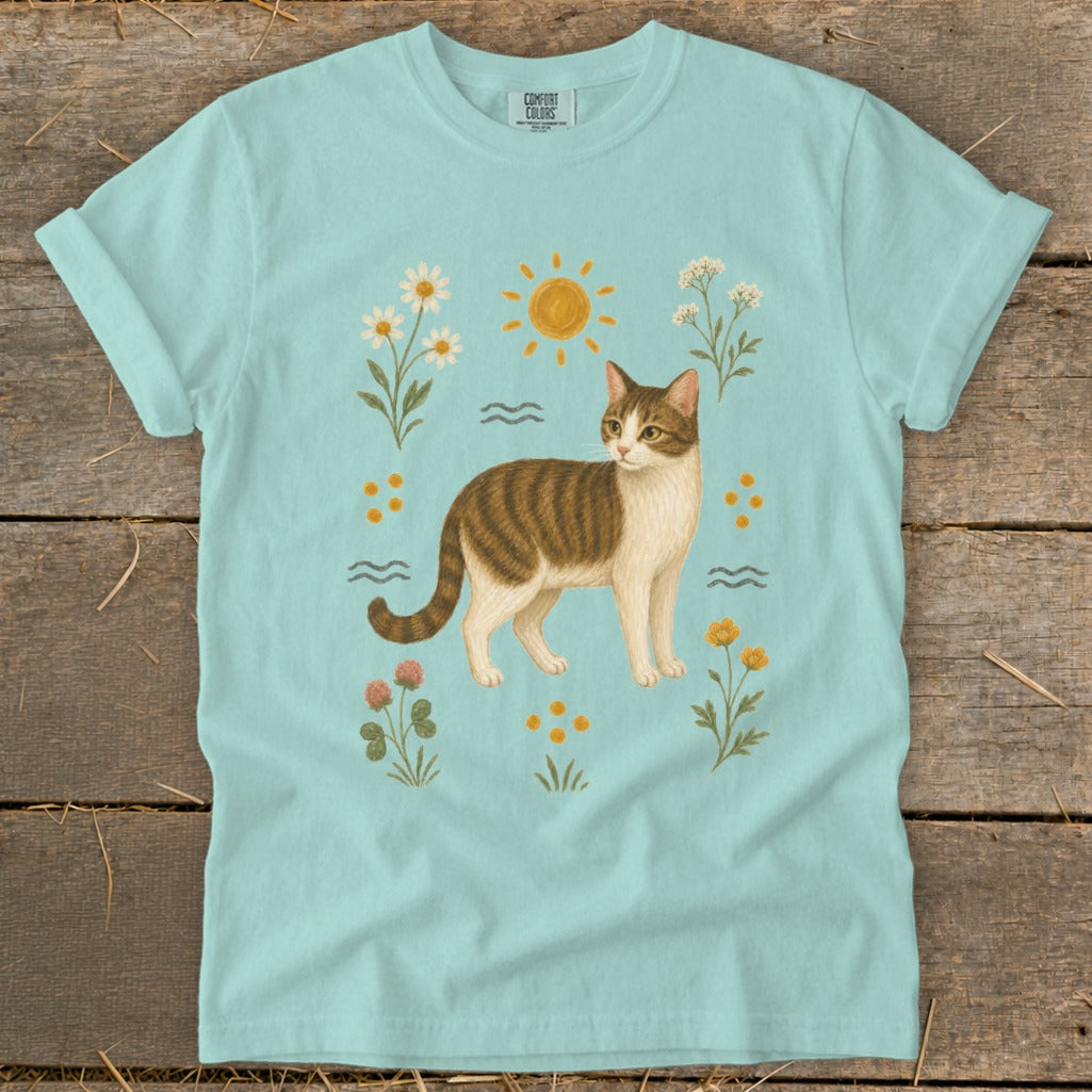 Light blue t-shirt with a brown and white tabby cat surrounded by flowers and sun.