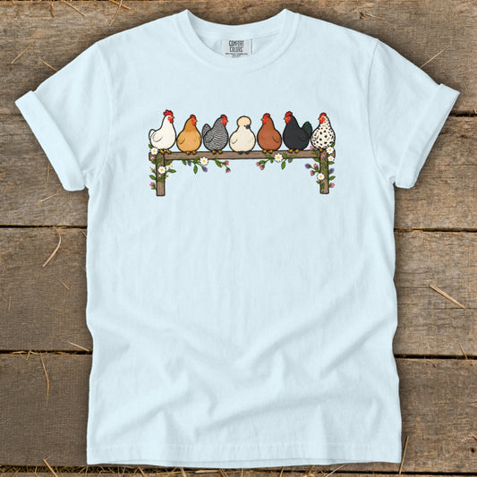 A light blue t-shirt with an illustration of seven chickens sitting on a wooden fence decorated with flowers.