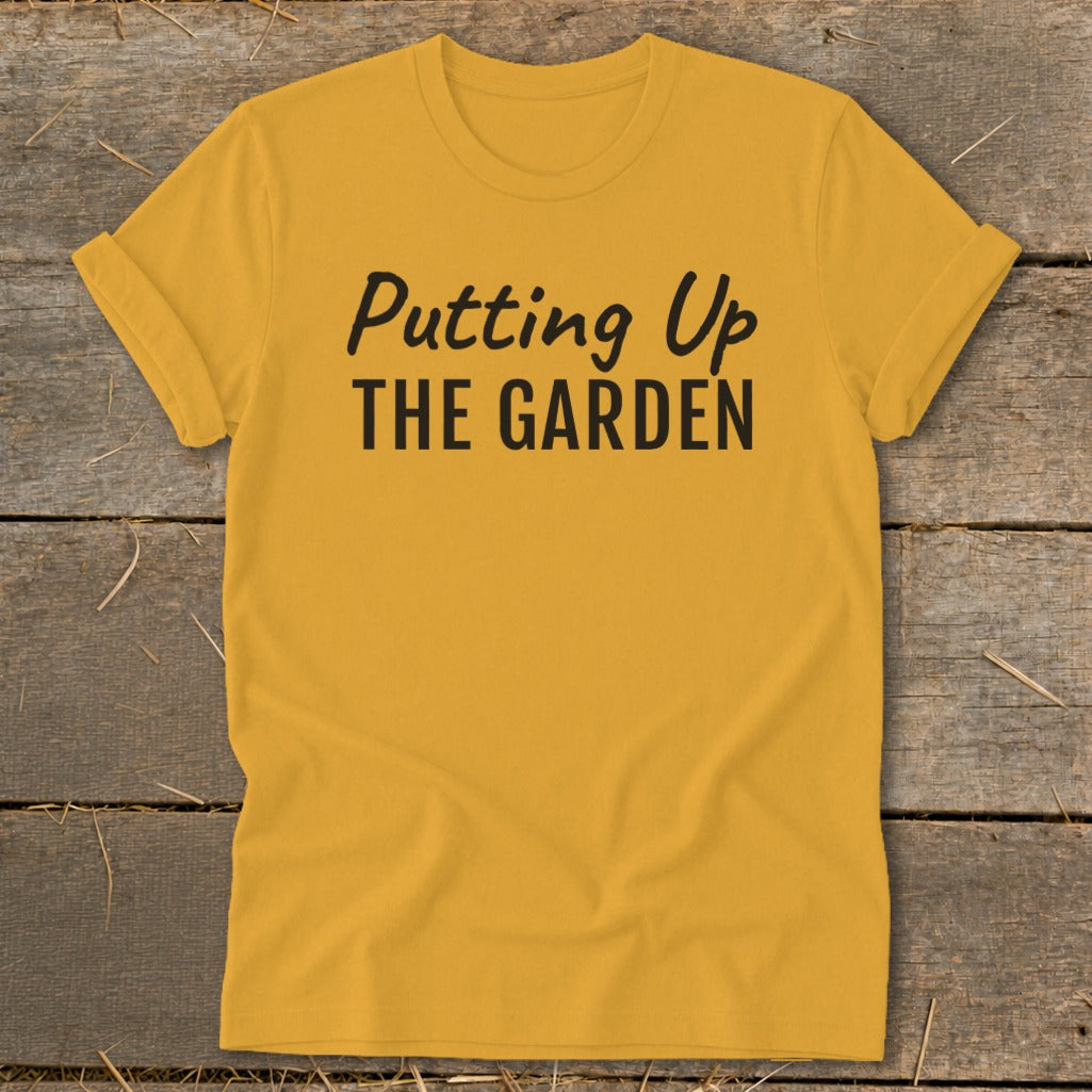 Yellow t-shirt with black text "Putting Up THE GARDEN" against a wooden background.
