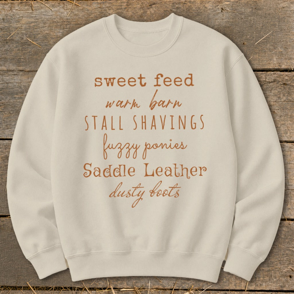 Cream sweatshirt with brown text listing equestrian terms: sweet feed, warm barn, stall shavings, fuzzy ponies, saddle leather, dusty boots.